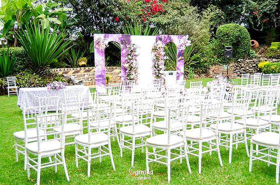 Rows of white chairs on the lawn facing a lilac and white floral arch, backed by tall garden planting