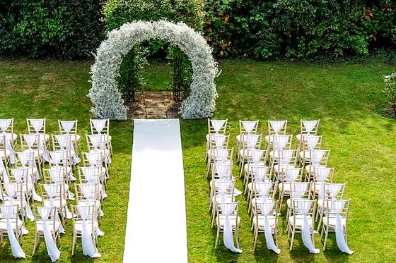 Tall white floral arch at the head of a white aisle runner with rows of chairs either side on the lawn