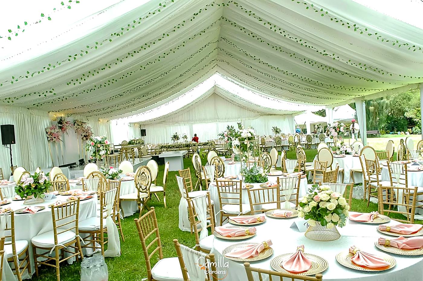 Marquee reception at Camilla Gardens with green garlands overhead, round tables laid in white and gold, and the garden visible through the open sides