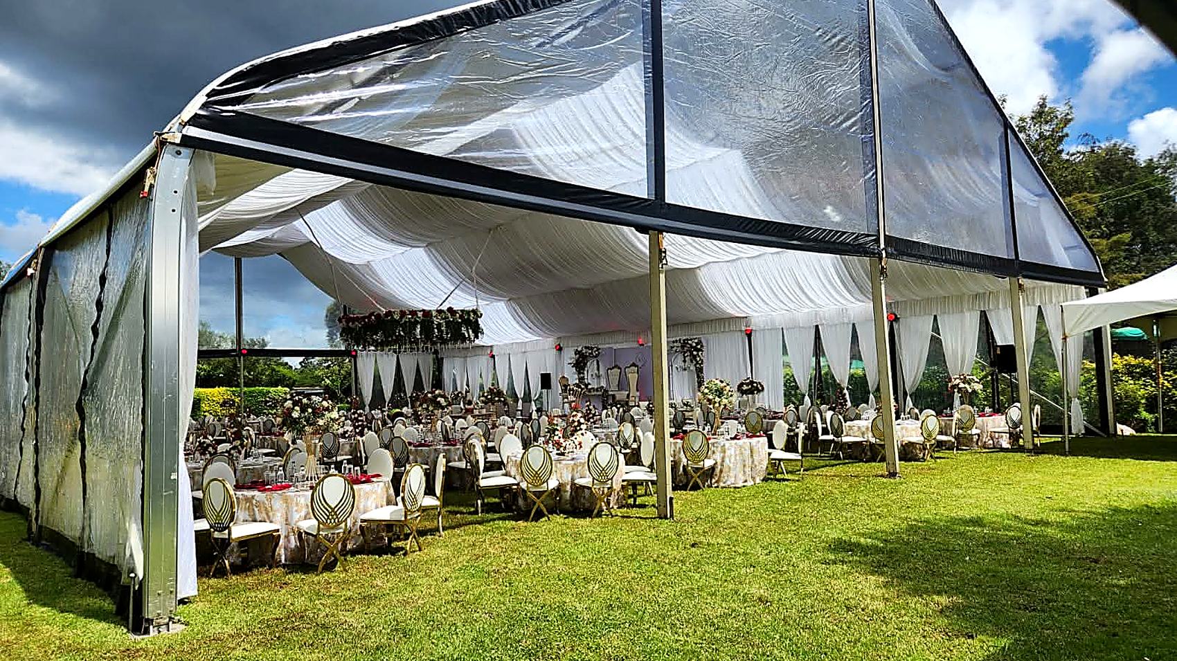 Clear-roofed marquee at Camilla Gardens standing open to the lawn, with round tables set beneath and garden trees behind