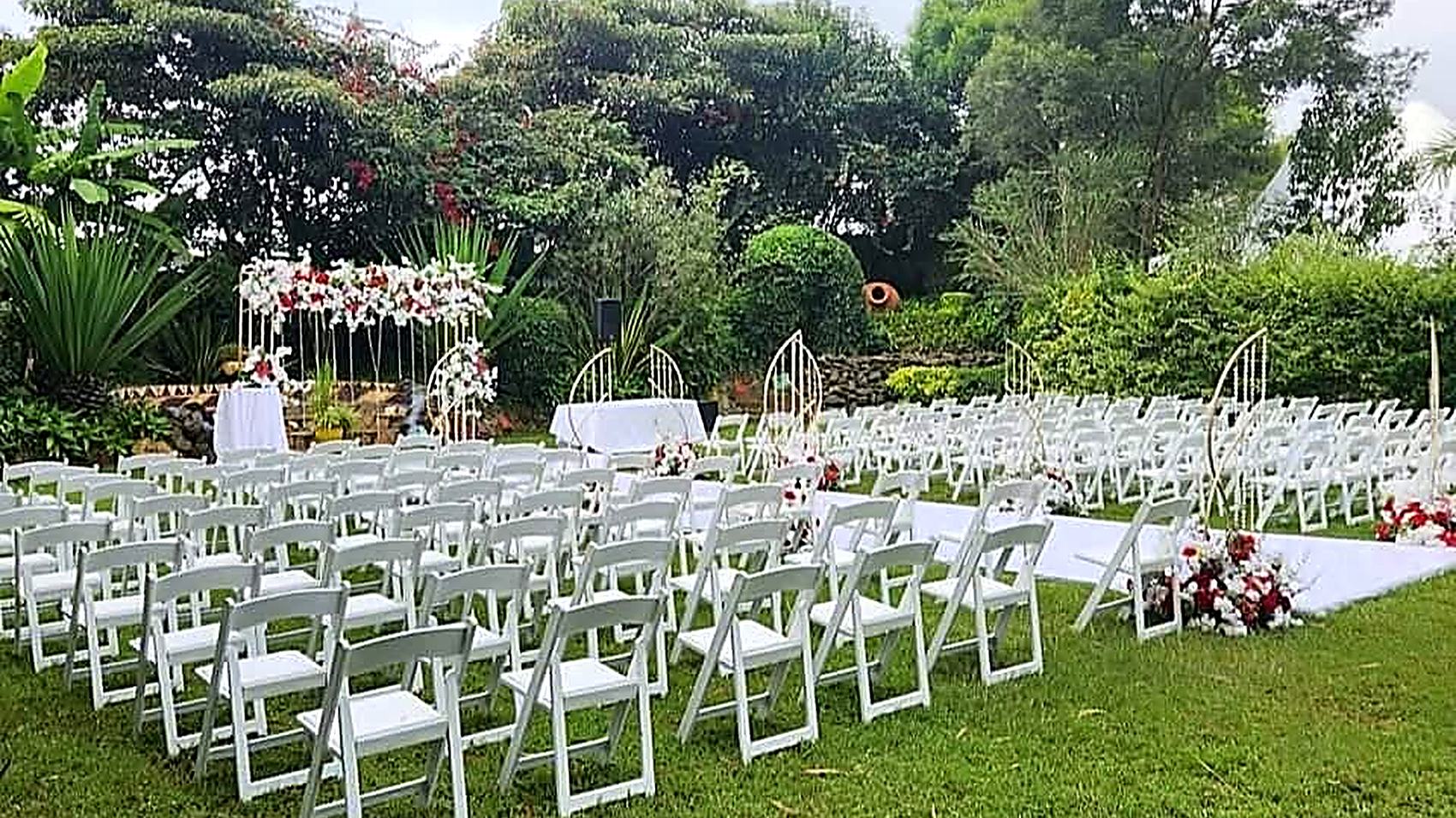 Church grounds at Camilla Gardens set for a ceremony, with a white-and-red floral arch and rows of white chairs facing a lawn backed by mature garden trees
