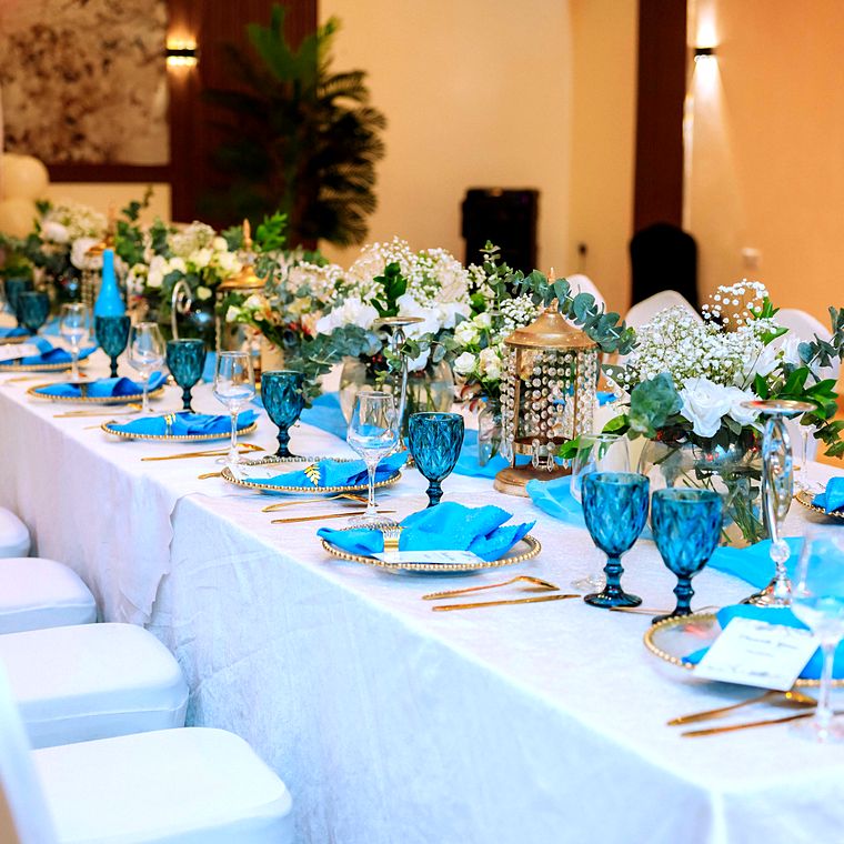 Long baby shower table with white cloth, sky-blue runners and napkins, teal goblets, gold beaded chargers and a crystal-drop lantern among white florals