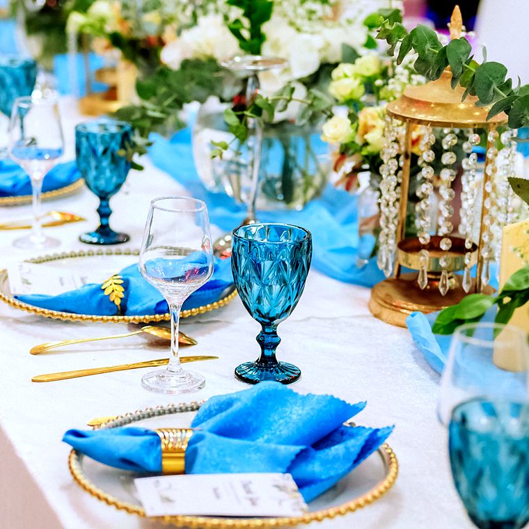 Close-up of a teal diamond-cut goblet and a clear wine glass beside a gold crystal-drop lantern, with blue napkins on gold beaded chargers
