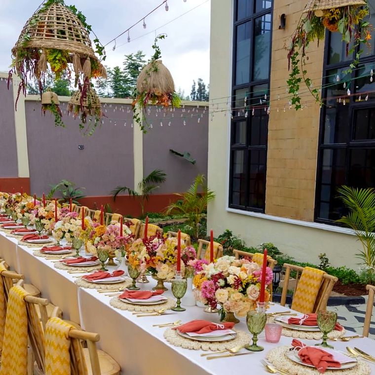 Courtyard long table under woven pendant planters trailing flowers, with coral napkins, red taper candles, green goblets and rattan placemats