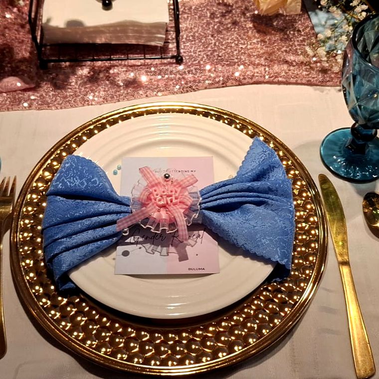 Top-down view of a place setting with a blue bow-folded napkin and a pink Baby Girl ribbon on a hammered gold charger