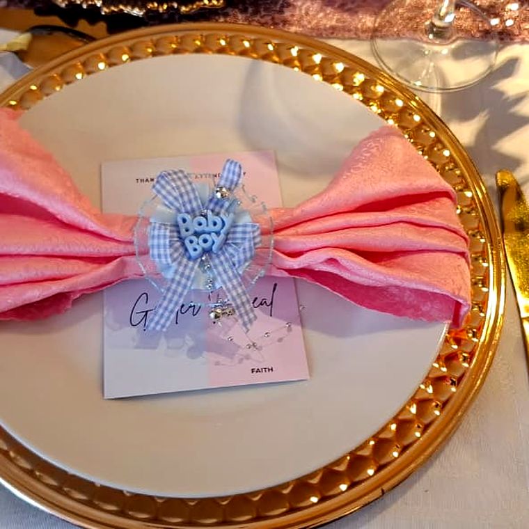 A pink bow-folded napkin with a blue Baby Boy ribbon on a gender reveal card, resting on a gold charger