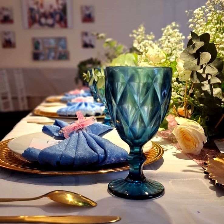 A teal diamond-cut goblet in front of a blue bow napkin on a gold charger, with white roses and eucalyptus behind