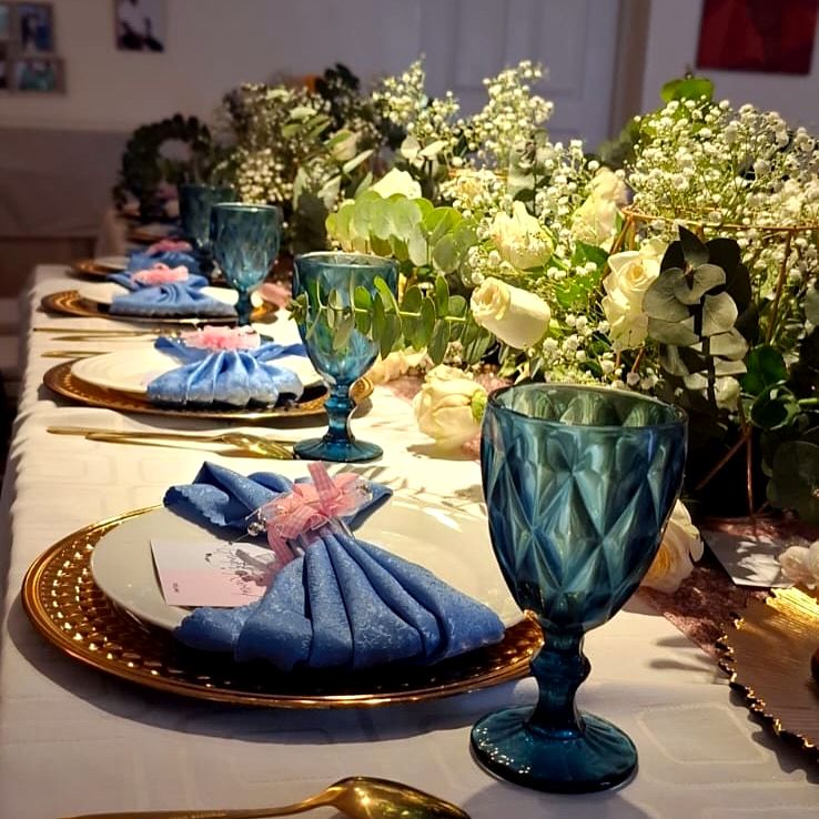 Gender reveal table with teal goblets, blue bow napkins on gold chargers, white roses, baby's breath and eucalyptus