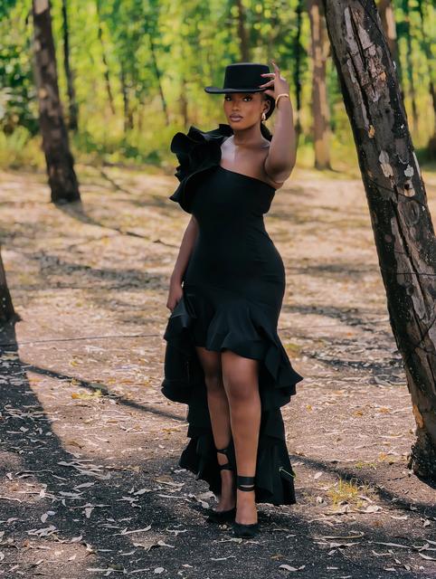 Woman in a one-shoulder ruffled black dress and flat-brim hat posing in a pine forest