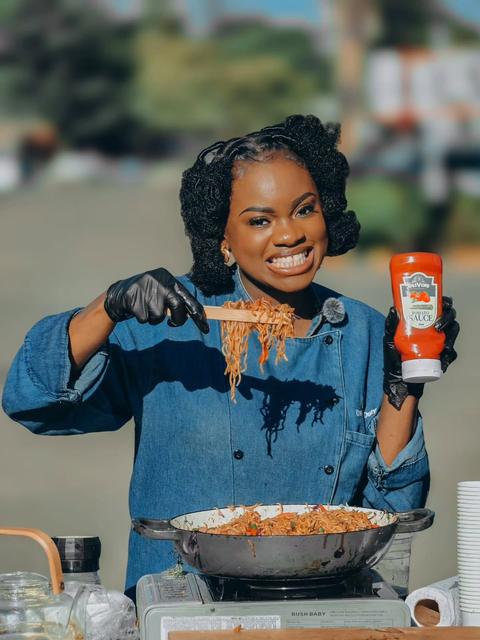 Chef On Duty in a denim chef jacket lifting a forkful of pasta from a wok, holding a sauce bottle and smiling