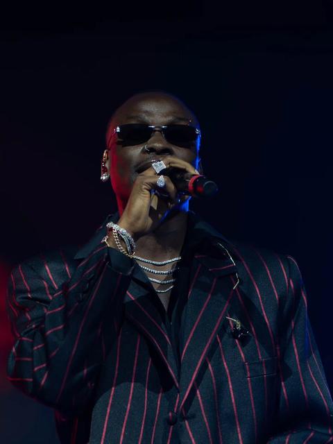 Fireboy DML performing on stage in a pinstripe suit and dark sunglasses, microphone to his mouth
