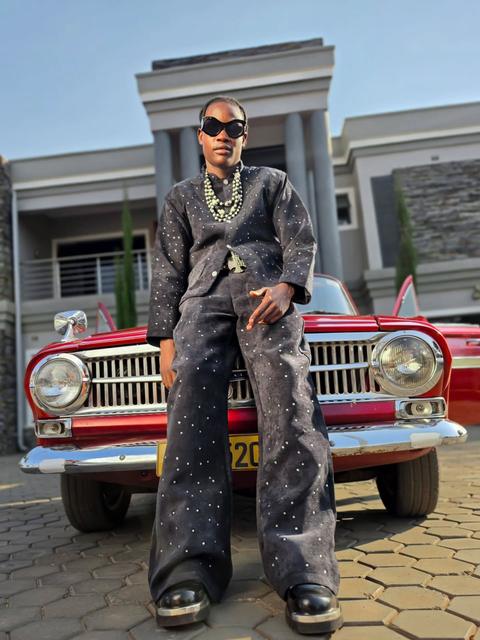 Man in a rhinestone-studded denim suit and sunglasses leaning on a red vintage car outside a mansion