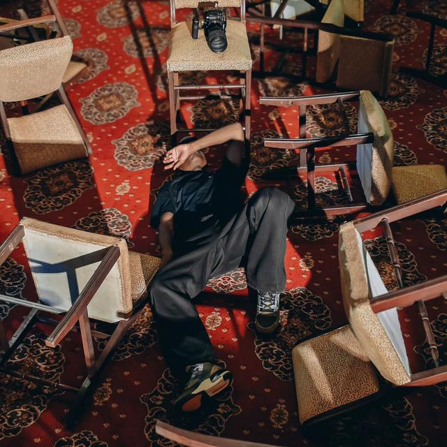 Carlos lying among overturned chairs on a red patterned carpet, arm over his face