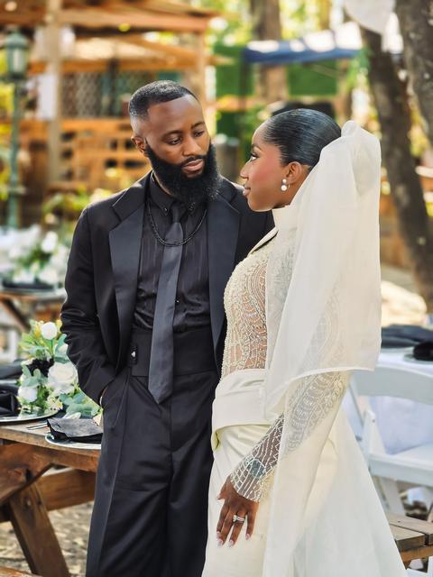 Groom in a black suit looking at his bride in a beaded high-neck gown and veil at an outdoor reception