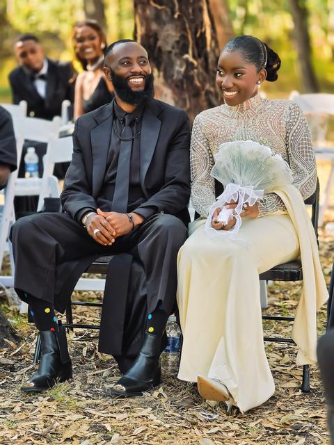 Bride and groom seated side by side, laughing, at an outdoor ceremony under trees