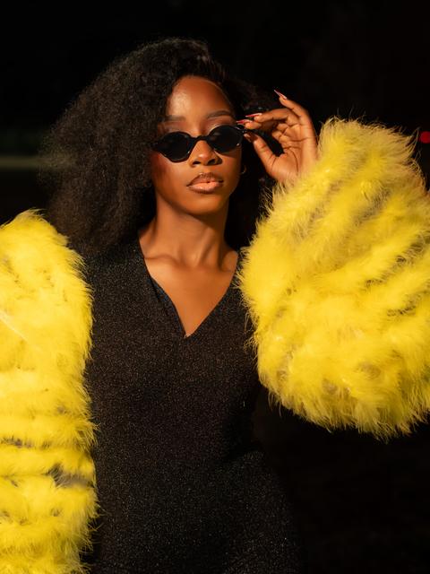 Woman in sunglasses with voluminous yellow feather sleeves, lit against a dark night background